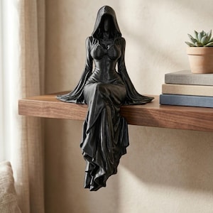 Pode incluir: Uma estatueta preta mate de uma figura encapuzada sentada com as pernas a balançar sobre uma borda. A figura veste uma túnica longa e fluida com mangas largas e um corpete justo. O capuz está levantado, ocultando o rosto da figura.