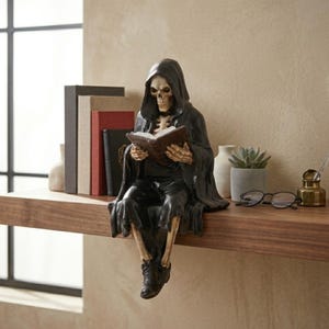 May include: A shelf-sitting figurine of a skeleton wearing a black hooded robe and reading an open book. The skeleton has a beige skull and ribcage, and wears black trousers and black shoes. The figure is positioned with legs dangling over the edge.