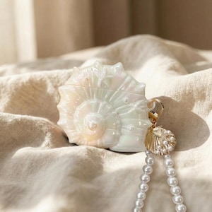 May include: A pearlescent white seashell-shaped case with a gold-colored charm and a strand of white pearls. The case has a spiral design and a glossy finish. The charm is a small seashell with pearl accents.