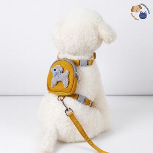 May include: Mustard yellow dog harness with a matching leash. The harness features a small backpack with a gray poodle applique and a blue bow tie. The harness and leash have gray plastic buckles and a silver metal clasp.