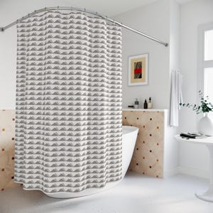 May include: A white shower curtain with a repeating pattern of gray and light gray triangles. The curtain hangs from a curved, silver-colored shower rod. The bathroom has a white bathtub and a small framed artwork.
