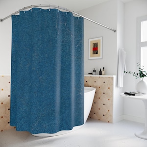 May include: A blue shower curtain with a textured appearance hangs from a curved, silver-colored rod. The curtain is the focal point in a bright bathroom setting, with a white bathtub and a small table visible.