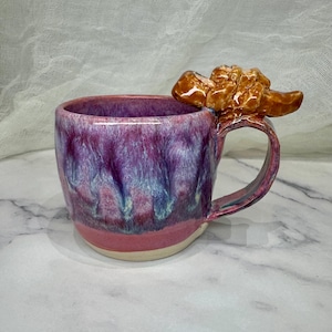 May include: A ceramic mug featuring a glossy glaze with blended shades of deep purple, magenta, and light blue. The mug has a unique handle topped with a sculpted, textured brown element that resembles a cluster of rocks or organic matter.