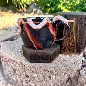 May include: A handmade ceramic mug with a dark brown base and red and black streaks. The mug features a handle and is adorned with pink and blue tentacle-like embellishments. It sits on a small wooden stand.