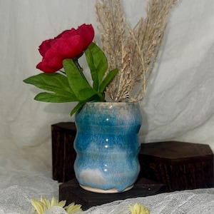 May include: A small, handmade ceramic vase with a wavy design, glazed in shades of blue and white. It holds a vibrant red flower with green leaves and dried ornamental grass. The vase is displayed on a dark wooden block.