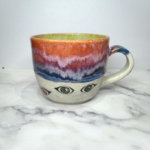 Può includere: Una tazza in ceramica con base bianco sporco maculata e occhi dipinti a mano in verde e rosa. La parte superiore presenta uno smalto vibrante a strati con una transizione dal blu scuro al viola, rosa e arancione, con interno verde chiaro.