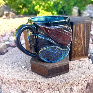 Pode incluir: Uma caneca de cerâmica artesanal com exterior preto, apresentando detalhes texturizados em vermelho e azul. A caneca tem um interior azul escuro e uma alça curva. Assenta num pequeno suporte de madeira escura, mostrando o design único e o artesanato.