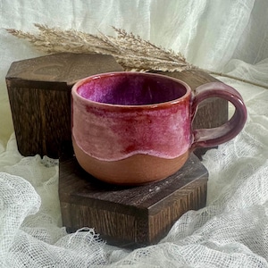 May include: A handmade ceramic mug with a burgundy interior and a reddish-pink exterior. The mug has a brown base and a curved handle. The mug is displayed on a dark wooden block.