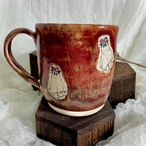 May include: A handmade ceramic mug with a red and brown glaze. The mug features three white ghost illustrations with floral accents. The mug is sitting on a dark wooden block.