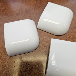 May include: Three white, square-shaped corner pieces with rounded edges are arranged on a brown surface. The pieces have a glossy finish, reflecting light. The objects appear to be components of a larger structure or product.