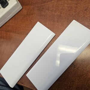 May include: Two rectangular white tiles with a glossy, smooth finish and beveled edges. The tiles are positioned diagonally, showcasing their reflective surface and clean, uniform appearance. Each tile features a simple, solid white color.