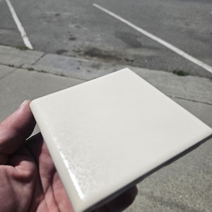 May include: A square, off-white ceramic tile with a smooth, glossy finish and rounded edges. The tile features a uniform, solid color surface that reflects light, highlighting its clean, minimalist design and polished, reflective texture.