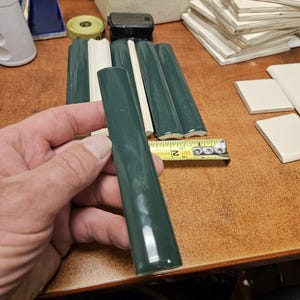 May include: A glossy dark green ceramic trim tile with a rounded edge profile. A yellow measuring tape is positioned next to the tile, showing a width of approximately 0.75 inches. The tile has a smooth, reflective surface and a uniform finish.