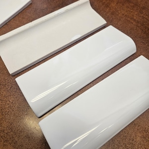 May include: Close-up of white ceramic tiles. The tiles are rectangular with a glossy finish. One tile has a curved edge. The tiles are arranged on a brown surface. The tiles are likely for a kitchen or bathroom.