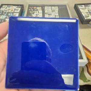 May include: A square, deep blue ceramic tile with a glossy, reflective surface. The tile features a smooth, solid color finish and a distinct, circular embossed spiral pattern visible on the right side of the surface, catching the light.