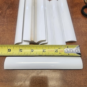 May include: A collection of white ceramic bullnose tiles with a glossy finish. A yellow Stanley measuring tape is positioned across the tiles, showing a width of approximately 6 inches for the individual tile piece placed in the foreground.