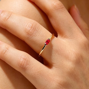 May include: A gold ring with a rectangular, bright red gemstone. The ring is simple in design, with a thin band and a single, eye-catching stone. The ring is shown on a finger, highlighting its delicate and elegant appearance.