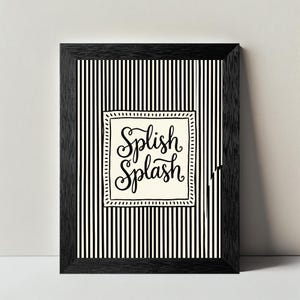 May include: A framed wall art print featuring a black and white vertical striped background. In the center, a cream-colored square with a dashed border contains the hand-lettered black text Splish Splash in a decorative cursive script font.
