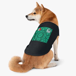 Puede incluir: Un perro Shiba Inu con una camiseta negra para mascotas. La camiseta presenta un diseño verde y blanco con la bandera y el emblema mexicanos. El perro es marrón y blanco, con la cola enroscada.
