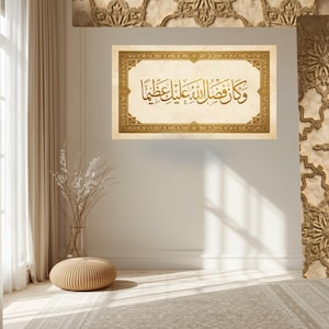 Peut inclure: Une œuvre d'art encadrée avec de la calligraphie arabe dorée sur fond beige. Le cadre présente une bordure décorative. L'œuvre est accrochée sur un mur blanc dans une pièce avec un tapis beige et un pouf tressé.