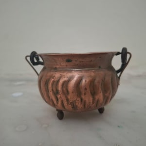 May include: A small, round copper pot featuring a textured, wavy vertical pattern on its exterior. The pot has two dark metal handles attached to the sides and rests on three small, rounded feet. The copper surface shows signs of natural patina.