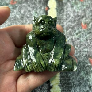 May include: A carved green stone monkey figurine. The monkey is sitting with its legs folded and arms resting on its knees. The figurine has detailed facial features and a textured surface.