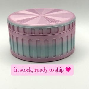 May include: A round, two-tone container with a lid. The lid and top portion of the container are pink with a radial design. The lower portion of the container fades from pink to teal. The text "in stock, ready to ship" is on a pink label.