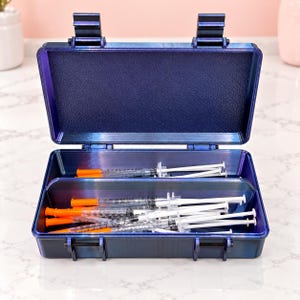 May include: A rectangular, dark blue, hard-shell storage case with a hinged lid, open to reveal two interior compartments. The case contains multiple clear plastic medical syringes with orange caps and white plungers arranged inside.