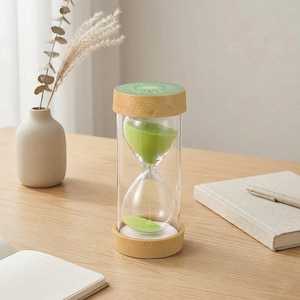 May include: A clear glass hourglass with a kiwi fruit design. The top and bottom are tan with a kiwi slice design on top. The sand inside is green. The hourglass is on a wooden table.