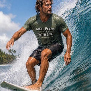 May include: A surfer rides a wave, wearing a green t-shirt with the text "MAKE PEACE WITH LIFE" and black shorts. The surfer is on a white surfboard, with water splashing around. The sky is blue with some clouds.
