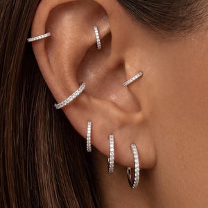 May include: A set of six silver-toned hoop earrings featuring small, clear, sparkling stones embedded along the outer surface. These circular earrings are positioned in various piercings across the ear, including the helix, rook, and lobe.