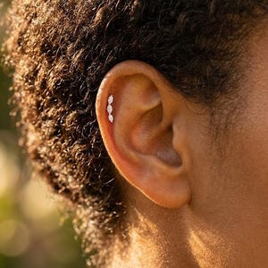 May include: Close-up of an ear featuring a triple marquise-cut diamond earring. The earring has a delicate, curved design, with three sparkling stones. The ear is set against a blurred background.