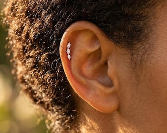 May include: A close-up of an ear with a triple marquise-cut diamond earring. The earring is set in a delicate, curved design, with three sparkling stones. The ear is set against a blurred background.