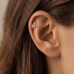 May include: A small gold-toned octopus-shaped stud earring featuring a polished finish and curved tentacles, positioned in the upper cartilage area of an ear. The jewelry piece has a symmetrical design with a rounded head and eight distinct limbs.