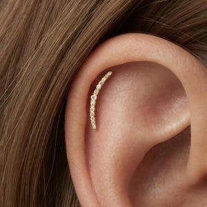 May include: A curved gold ear cartilage earring featuring a row of small, clear, round-cut stones set along the entire length of the arc. The piece is positioned vertically along the upper ear cartilage, showcasing a delicate and shimmering design.