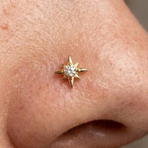 May include: A gold nose stud featuring a four-pointed star design with a central clear gemstone. The star has elongated, sharp points extending from the center, creating a celestial shape that sits flat against the skin surface.