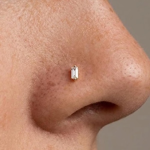 May include: A gold nose stud featuring a single rectangular baguette-cut clear gemstone set vertically. The jewelry piece is secured in the nostril, showcasing a polished metal setting that holds the stone in a clean, linear orientation.