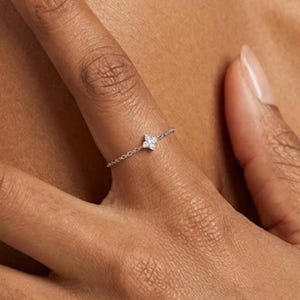 Op de afbeelding: Een delicate zilveren ring met een vierbladige bloemvorm, bezet met kleine, sprankelende edelstenen. De ring wordt aan een vinger gedragen en benadrukt de ingewikkelde details en elegante eenvoud. De kettingachtige band van de ring voegt een unieke touch toe.
