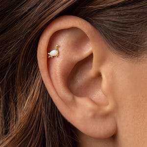 Può includere: Un orecchino a lobo in oro a forma di dinosauro dal collo lungo, caratterizzato da un inserto in opale bianco iridescente e liscio per il corpo. Il piccolo e delicato gioiello è posizionato sulla cartilagine superiore dell'orecchio con una finitura lucida.