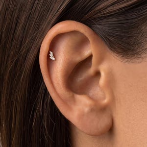 May include: A small, metallic ear piercing stud featuring three clear, marquise-cut stones arranged in a vertical, slightly curved line. The jewellery is positioned in the upper cartilage area of the ear, showcasing a delicate, elongated design.