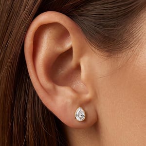 May include: A gold stud earring featuring a pear-shaped clear gemstone set in a smooth bezel setting. The earring is positioned in the earlobe, showcasing the teardrop silhouette of the stone and the polished yellow gold metal frame surrounding it.