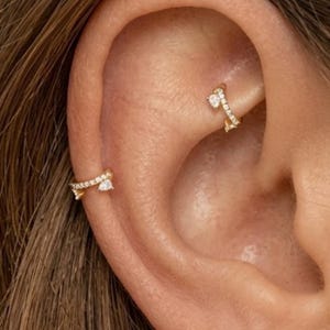 May include: Two gold-toned ear cuffs worn on the cartilage and helix of an ear. Each piece features a thin, curved metal band encrusted with small, clear, sparkling stones and a single, prominent pear-shaped stone at one end of the opening.
