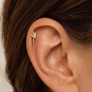 Könnte beinhalten: Ein goldfarbener Ohrstecker in Form einer stilisierten Libelle mit zarten, durchbrochenen Flügeln und einem schlanken Körper, platziert am oberen Knorpel des Ohrs. Das Schmuckstück hat eine polierte Metalloberfläche und einen Stecker.