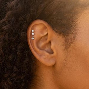 May include: Close-up of an ear adorned with a triple-stone earring. The earring showcases three clear, round gemstones, set in silver or white metal. The gemstones are linked by small chains, creating a dangling effect.