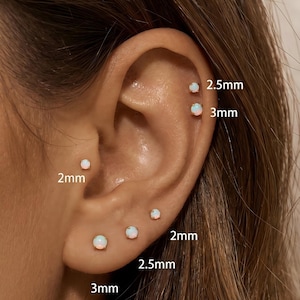 May include: A set of six round opal stud earrings in gold settings, displayed in various ear piercings. The studs are labeled with sizes 2mm, 2.5mm, and 3mm, indicating the diameter of each opal stone. The earrings feature a classic prong setting.