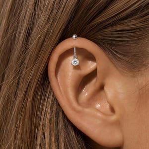Könnte beinhalten: Ein silberner Industrial-Barbell-Ohrring mit einem kleinen, hängenden, runden, klaren Edelstein. Der Ohrring wird in einem Ohrpiercing gezeigt. Der Barbell hat an einem Ende eine kleine Kugel, an der anderen der Edelstein hängt.
