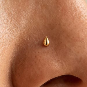 Può includere: Un piccolo piercing al naso in oro lucido a forma di goccia liscia e arrotondata. Il gioiello presenta una superficie metallica riflettente ed è posizionato centralmente sulla pelle, mostrando una forma semplice senza pietre aggiunte.
