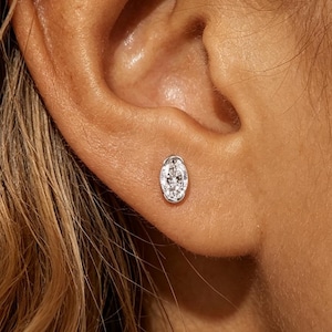 May include: A silver-toned stud earring featuring an oval-shaped setting with a cluster of small, clear, sparkling stones embedded within the center. The earring is positioned securely in the earlobe, showcasing a polished metallic finish.