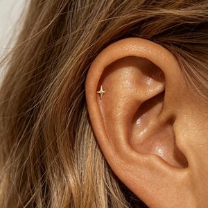 Könnte beinhalten: Ein kleiner goldener Ohrstecker in Form eines vierzackigen Sterns, der im oberen Knorpelbereich eines Ohrs sitzt. Der Ohrstecker hat eine polierte metallische Oberfläche mit scharfen, symmetrischen Spitzen, die von einem Zentrum ausgehen.