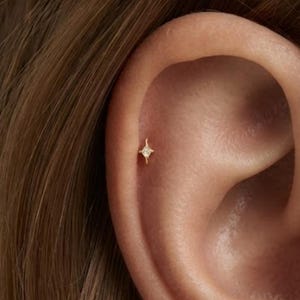 Könnte beinhalten: Ein kleiner goldfarbener Ohrstecker in Form eines vierzackigen Sterns mit einem zentralen, klaren Stein. Der Ohrstecker befindet sich im oberen Knorpelbereich eines Ohrs und zeigt ein filigranes Design mit dünnen, spitz zulaufenden Strahlen.